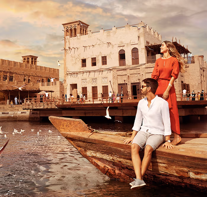Couple on a traditional wooden boat (abra) near the Al Seef heritage buildings, surrounded by flying seagulls at sunset.
