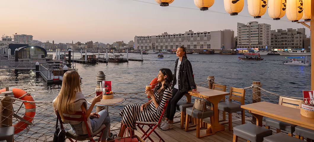 Three women enjoy drinks and conversation at an outdoor waterfront café at sunset, with lanterns overhead and boats visible on the water in the background.