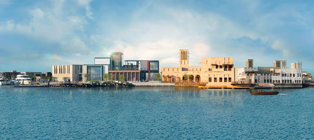 A scenic waterfront view featuring a mix of modern and traditional Middle Eastern architecture, with boats docked along the shore and a small boat sailing on the water under a blue sky.