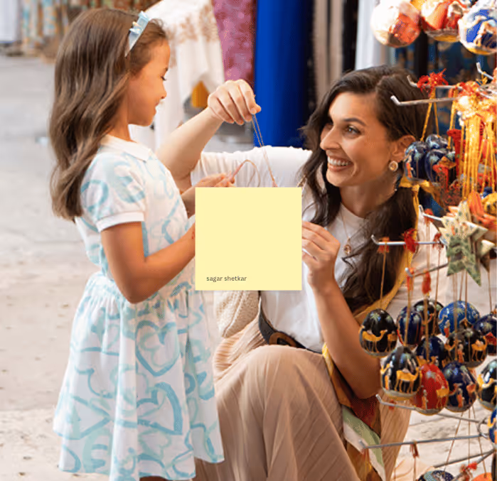 Smiling mother and daughter looking at hanging handcrafted ornaments at a festive market stall, full of colorful decorative items.