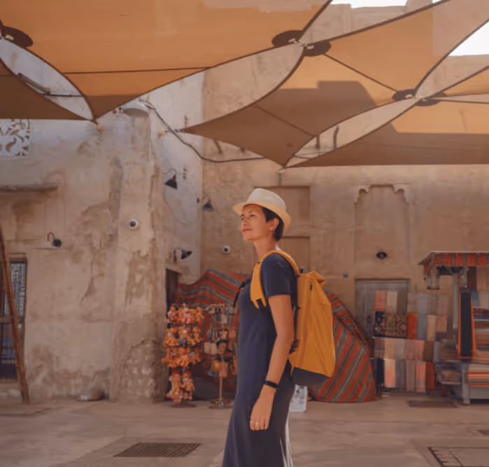 Female traveler with a yellow backpack walking through a shaded area of an old souk, surrounded by stone walls and colorful textiles.