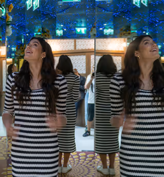 A woman in a black-and-white striped dress stands in a mirrored room decorated with flowers and twinkling blue lights, smiling and looking upwards as her reflection repeats around her.