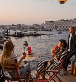 Three women enjoy drinks and conversation at an outdoor waterfront café at sunset, with lanterns overhead and boats visible on the water in the background.