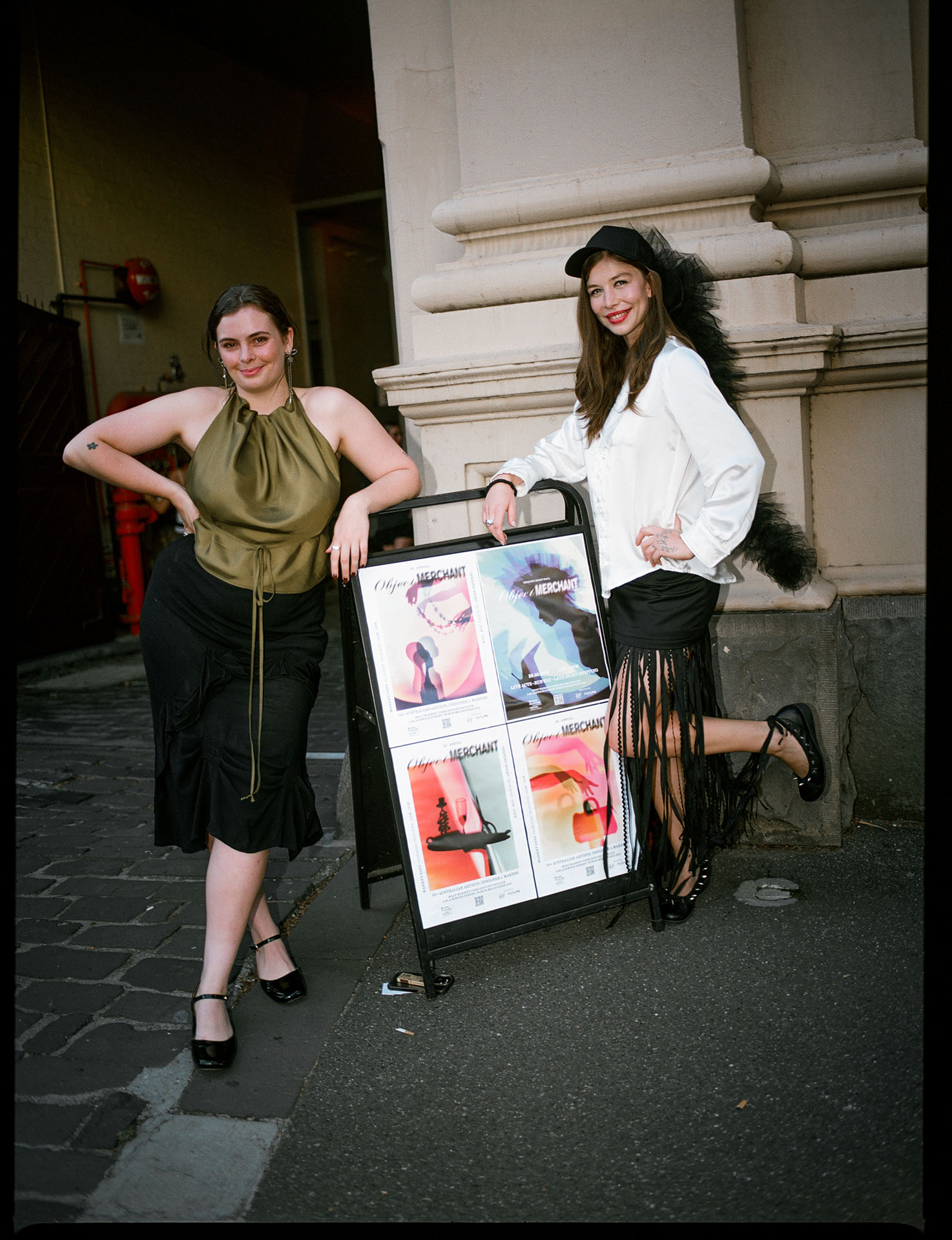 Image of two women leaning against a large sign, outside of a dramatic entryway