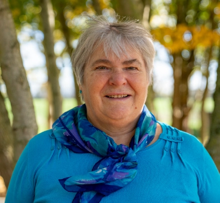 Smiling older woman with short gray hair wearing a blue top and a blue patterned scarf, standing outdoors with trees in the background.