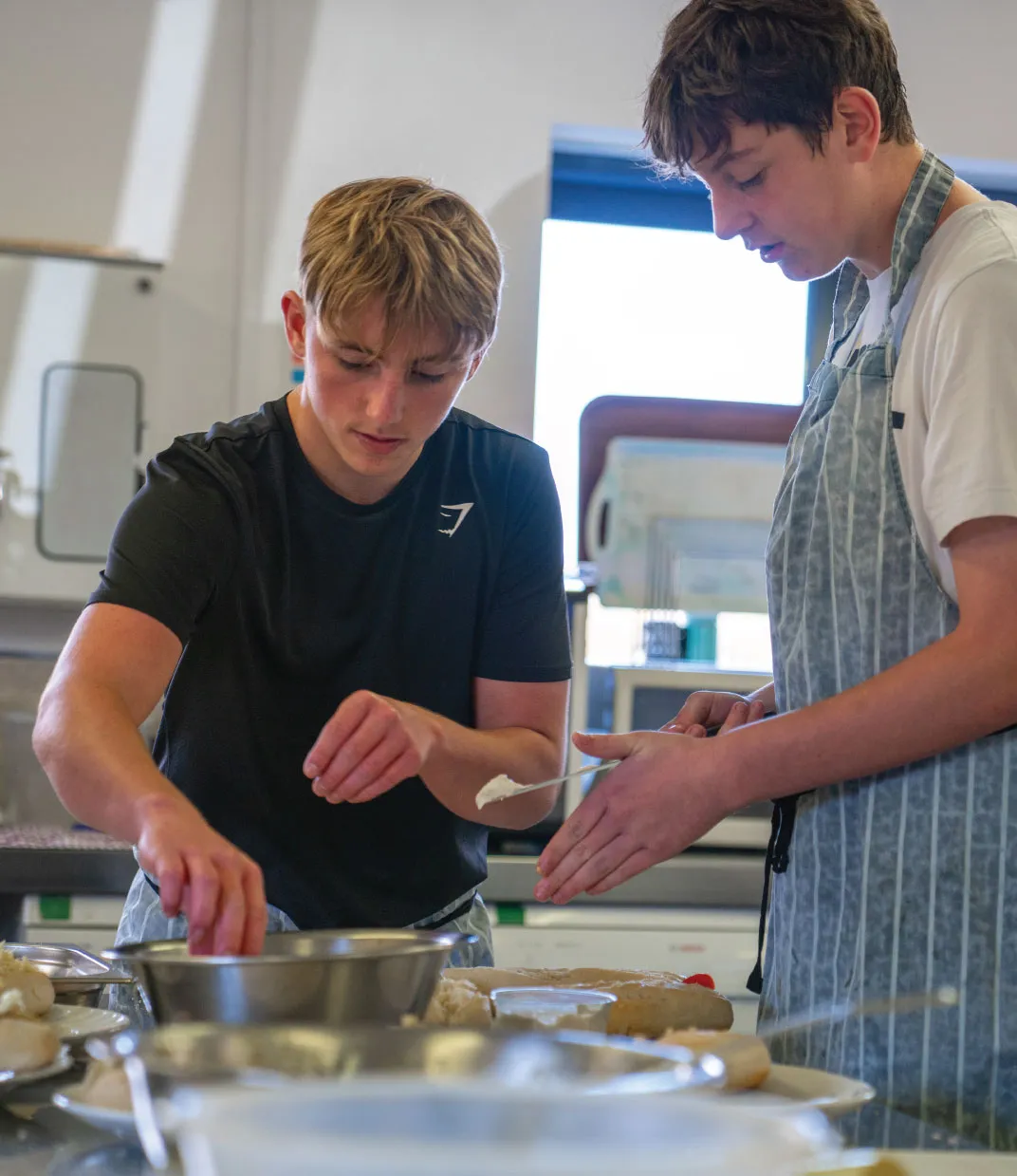 Two teenage boys cooking together in a kitchen, one wearing a black shirt and the other a striped apron, preparing food with bowls and utensils on the counter.