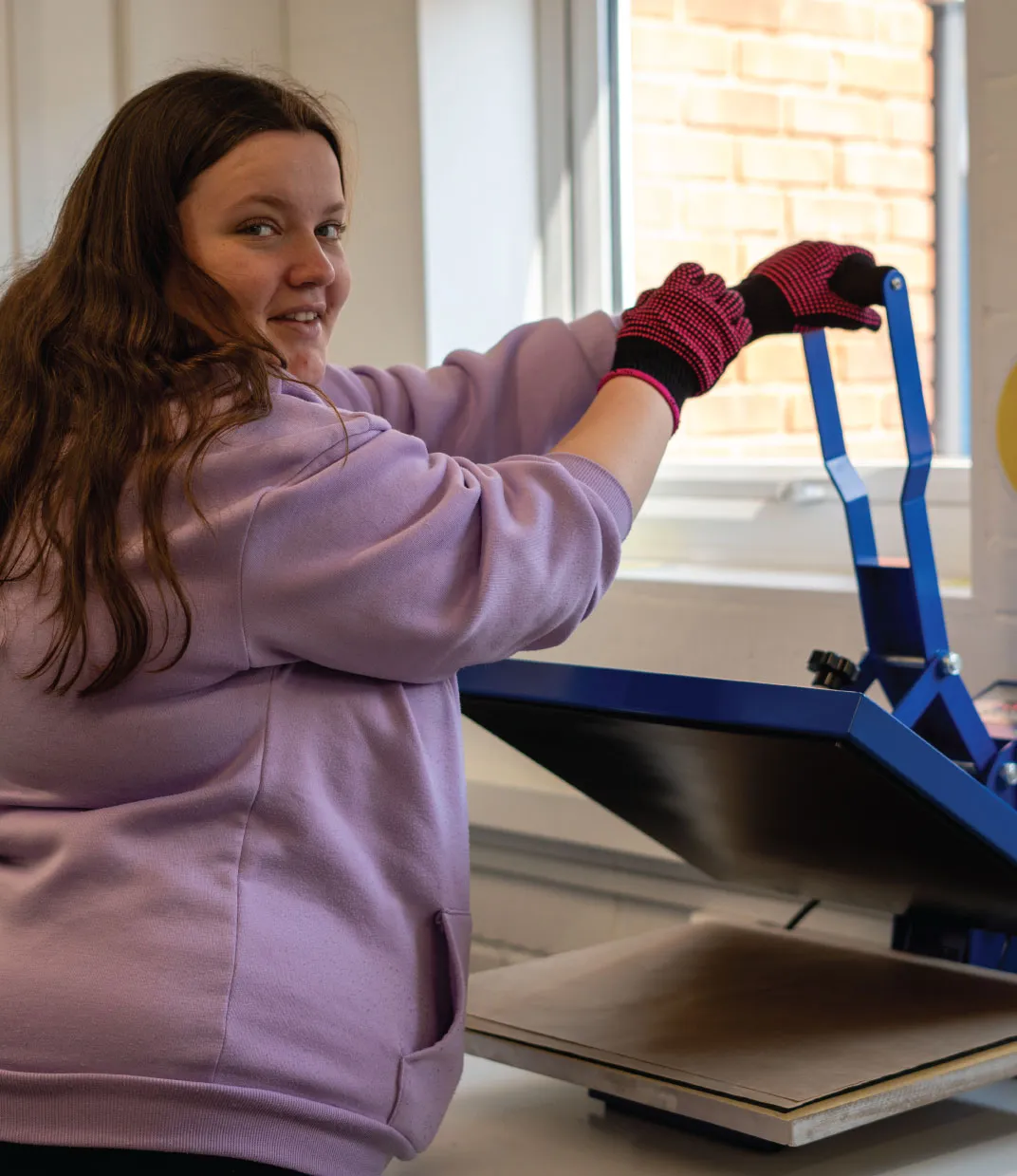 Person in purple sweatshirt and black gloves operating a blue heat press machine near a window.