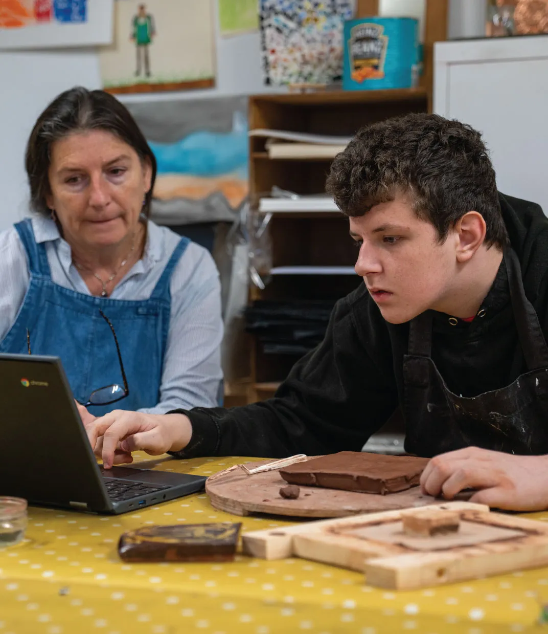 Teacher and student working together on a laptop with clay art materials on the table.