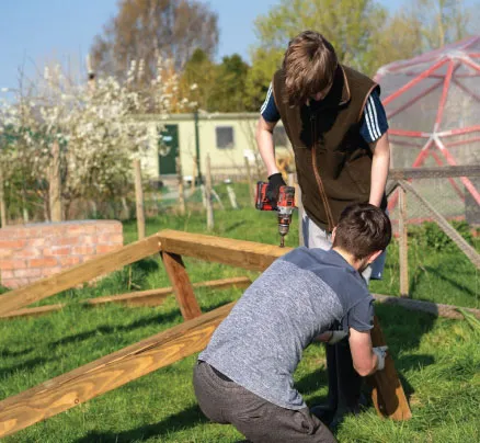 Two young students outdoors assembling a wooden frame, one using a drill while the other holds the wood steady.