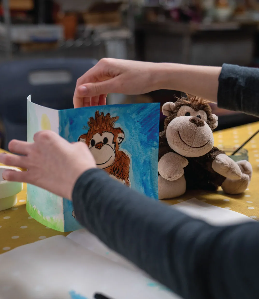 Person holding a hand-painted card with a smiling monkey drawing next to a stuffed monkey toy on a yellow polka dot tablecloth.