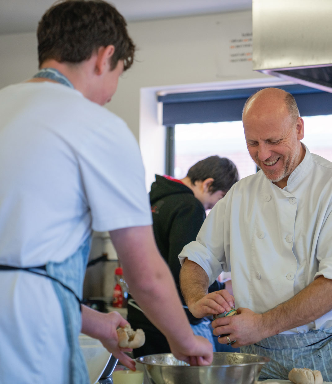 Smiling chef in white uniform preparing food with two younger people in a kitchen.