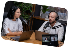 Two people wearing headphones speaking into microphones during a podcast recording with a laptop and screen showing remote participants.