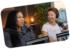 Two women speaking into microphones in a podcast studio with acoustic panels and plants.