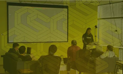 Group of five people in a casual meeting room with one standing and presenting from a laptop while others sit and listen.