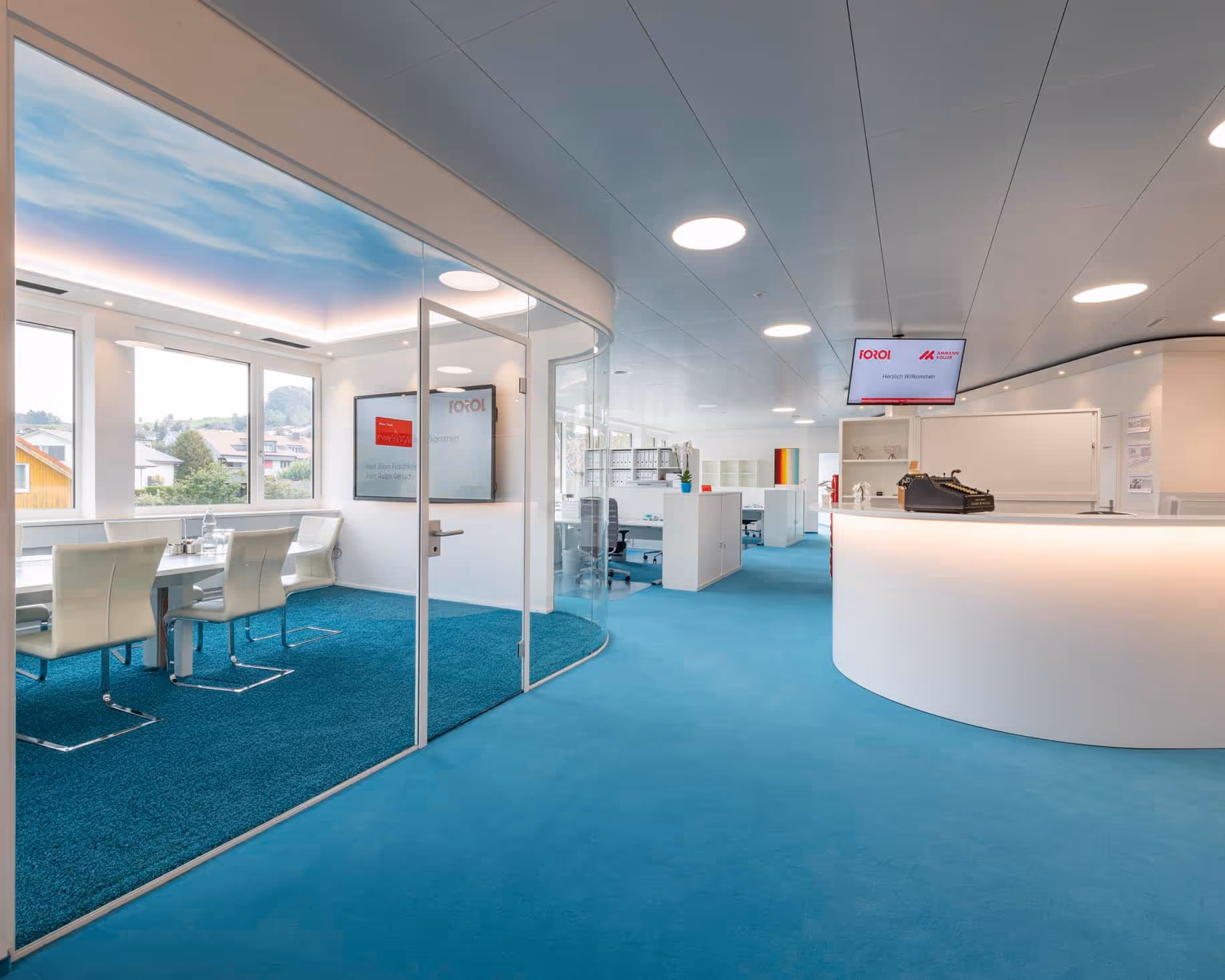 Modern office interior with turquoise carpet, glass-walled meeting room with white chairs, and a curved white reception desk.
