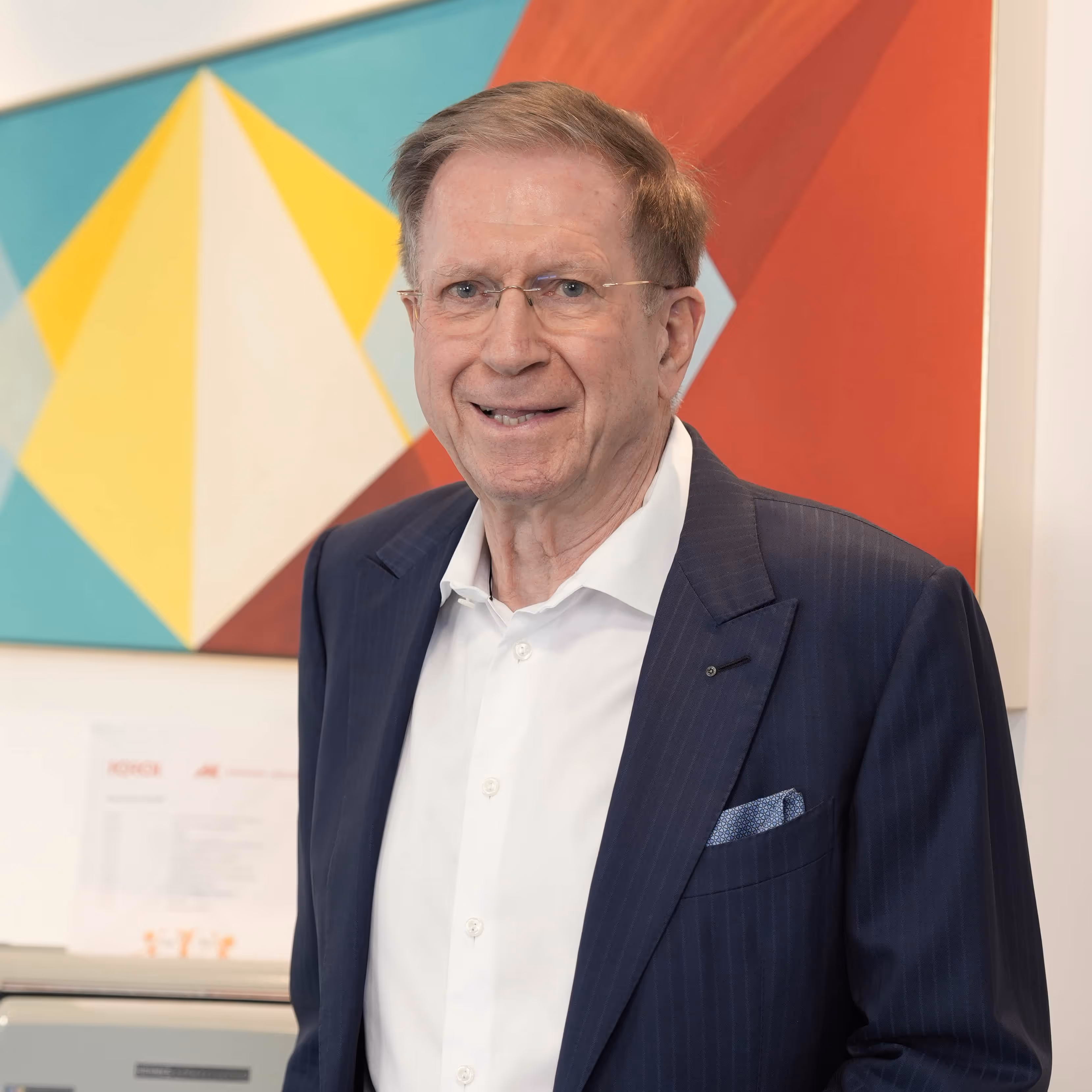 Older man wearing glasses and a navy suit jacket with a white shirt, standing in front of colorful abstract art.