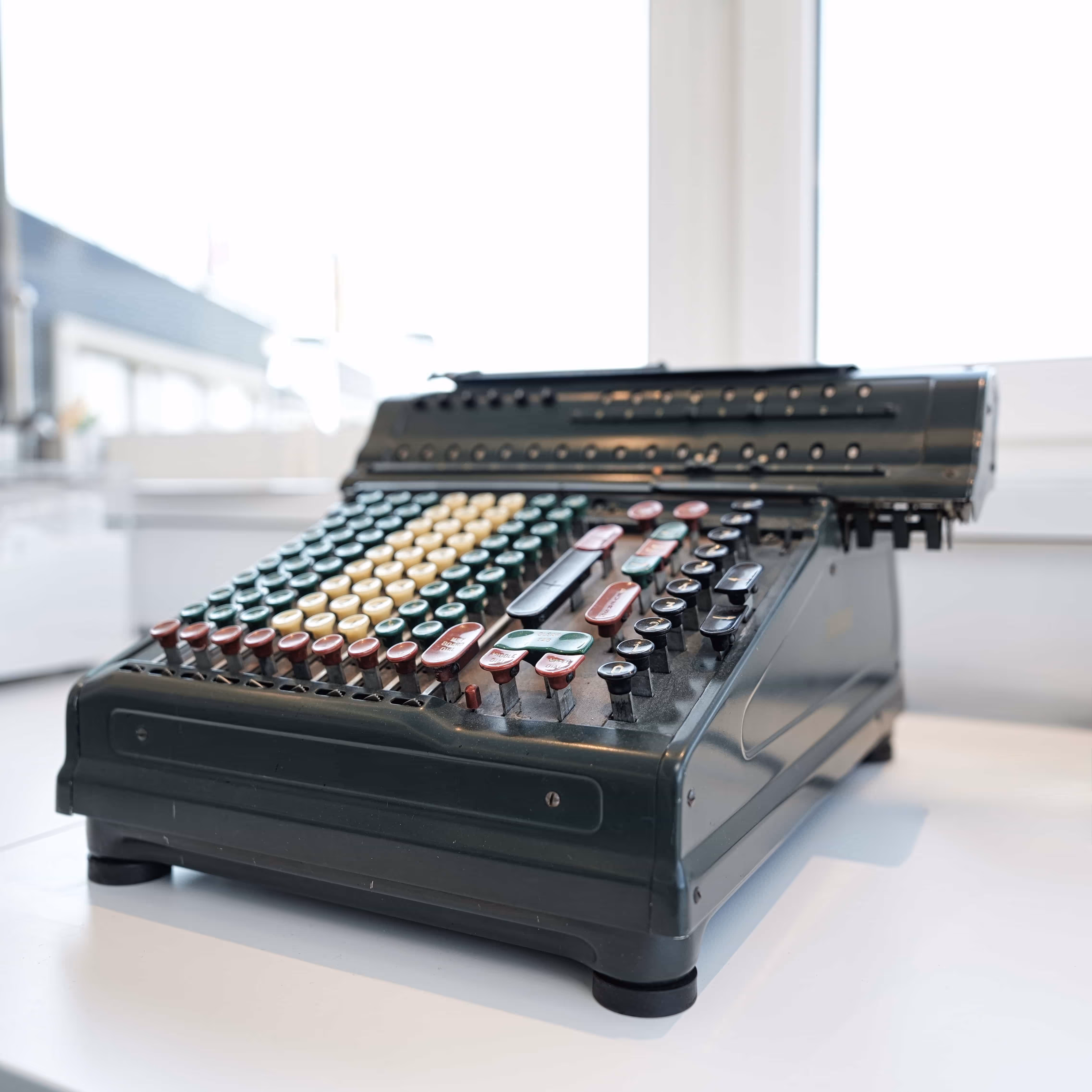 Vintage mechanical cash register with multiple round and rectangular buttons in red, green, cream, and black on a white surface near a window.
