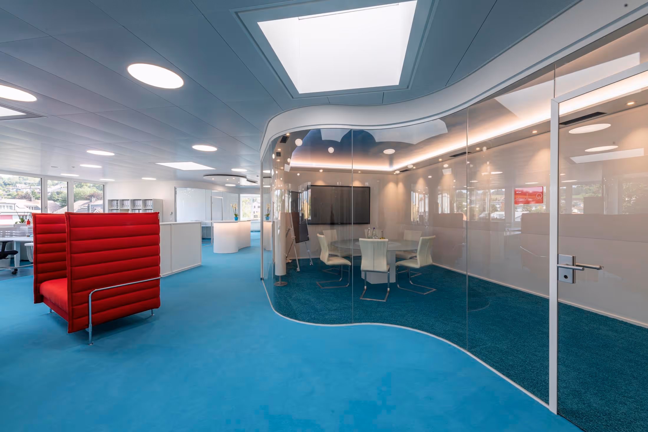 Modern office space with blue carpet, curved glass-walled meeting room with white chairs and round table, and a large red cushioned bench.
