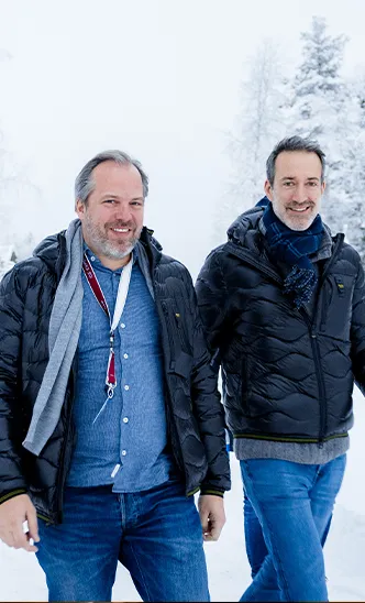Zwei lächelnde Teilnehmer des ESC European Speed Club in Winterjacken bei einem Event in einer verschneiten Landschaft.

