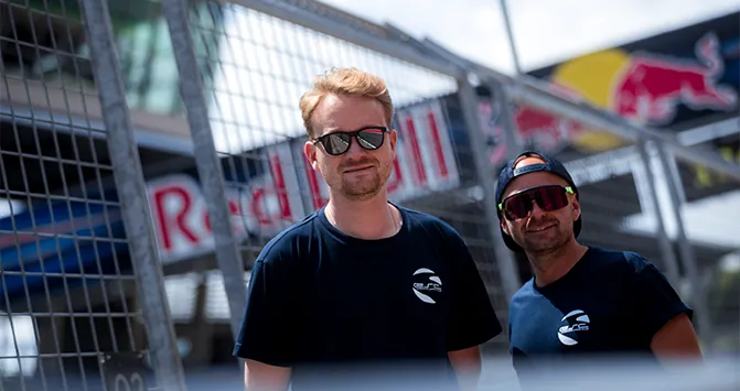 Zwei Mitglieder des ESC European Speed Club in Team-Shirts posieren lässig vor der Red Bull-Kulisse auf der Rennstrecke.

