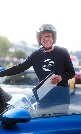 Ein freundlicher Instruktor des ESC European Speed Club mit Helm und Klemmbrett steht neben einem Auto auf der Rennstrecke.