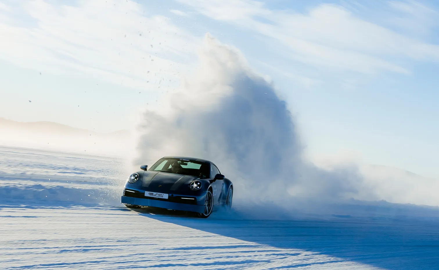 Blauer Sportwagen driftet auf verschneiter Fläche und wirbelt eine große Schneewolke auf.