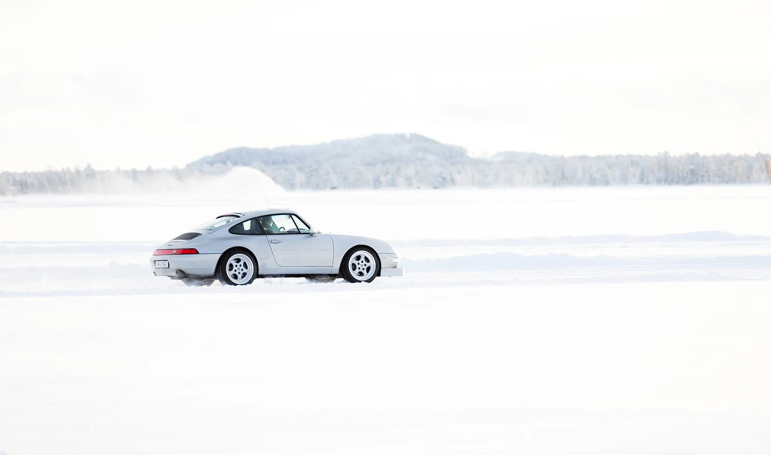 A classic silver Porsche 993 from the ESC European Speed Club drifts across a frozen lake in Sweden.

