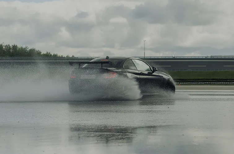 Schwarzes Sportcoupé fährt auf nasser Fahrbahn und spritzt Wasser seitlich auf.