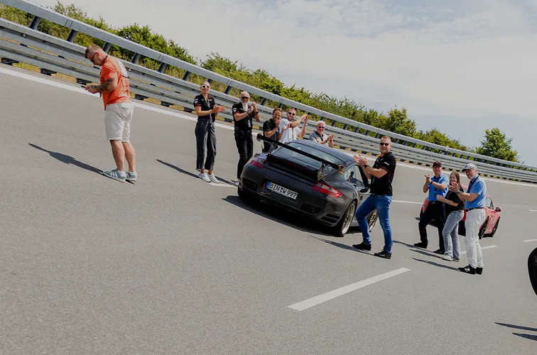 Gruppe von Menschen applaudiert neben einem schwarzen Sportwagen auf einer kurvigen Straße mit Metallgeländer.