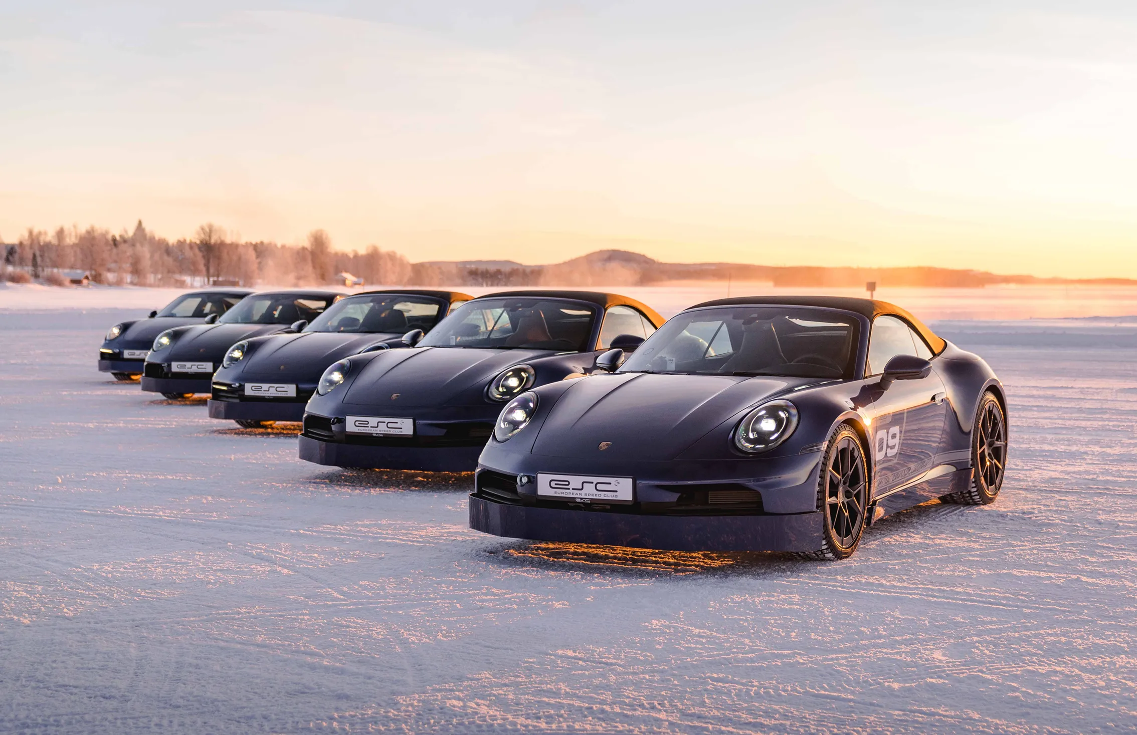 Fünf dunkle Porsche-Cabriolets, in einer Reihe auf einer schneebedeckten Fläche bei Sonnenuntergang geparkt.