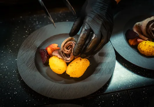 Zwei gelbe Kroketten und ein Stück gerolltes Fleisch werden auf einem schwarzen Teller mit schwarzen Handschuhen angerichtet.