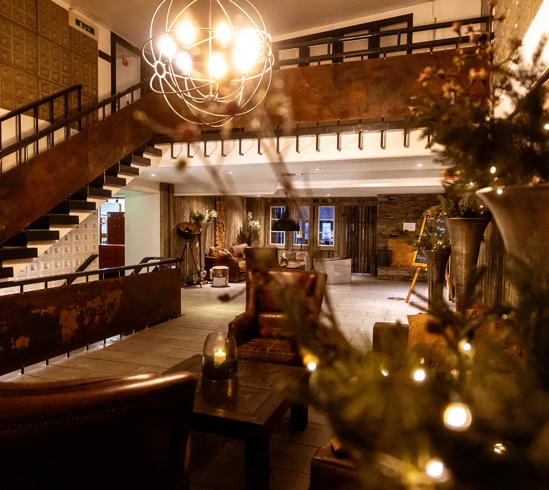 Gemütlicher, warm beleuchteter Hotel-Lobbybereich mit Ledersesseln, Holztischen und einer Treppe mit Metallgeländer.