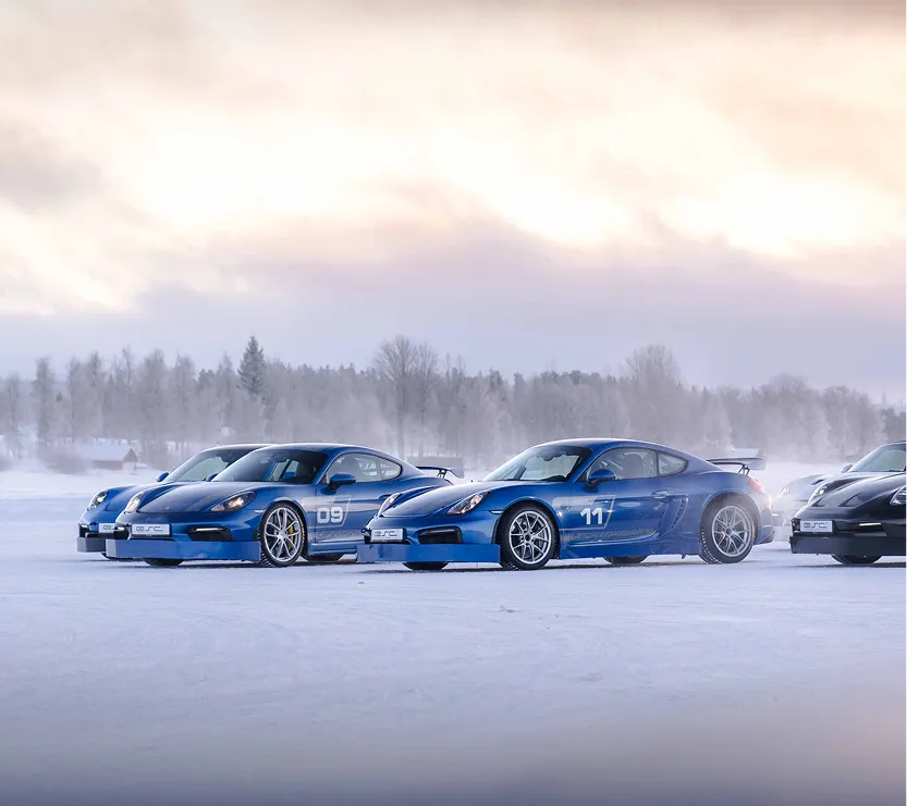 Drei blaue Sportwagen mit Rennnummern stehen auf verschneiter Fläche vor nebligem Wald.