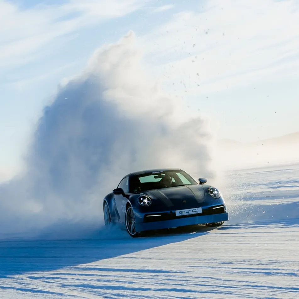Blauer Sportwagen driftet auf verschneiter Fläche, wirbelt eine große Schneewolke auf.