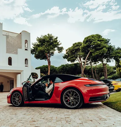 Roter Porsche 911 Cabrio auf gepflasterter Einfahrt vor einem Haus mit Bäumen im Hintergrund.
