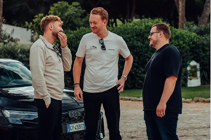 Drei Männer stehen draußen vor einem schwarzen Auto und unterhalten sich lachend.