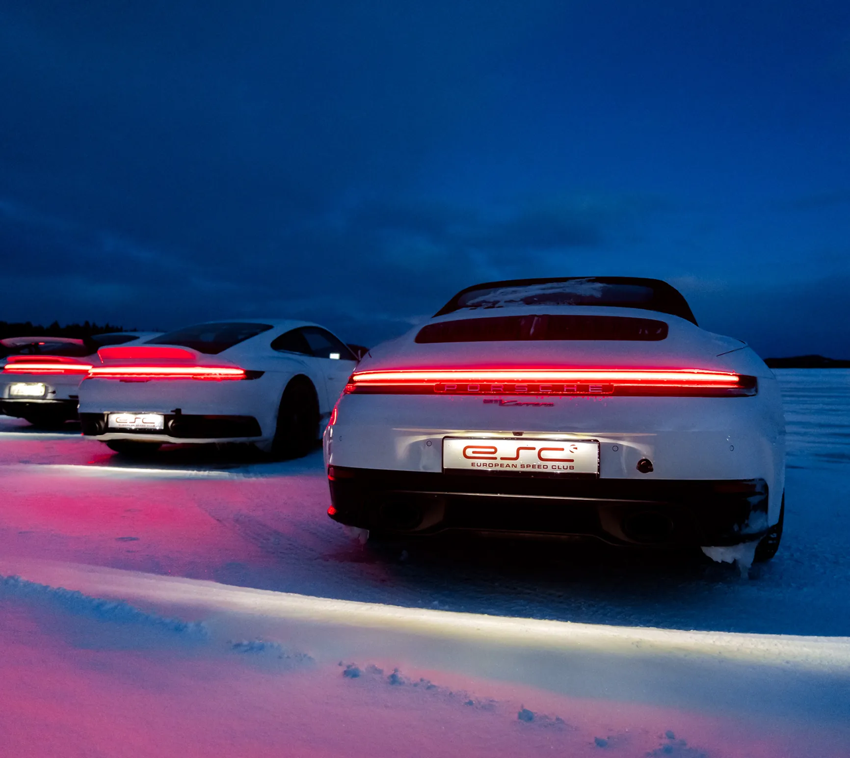 Weiße Porsche-Sportwagen mit leuchtenden Rücklichtern stehen bei Dämmerung auf verschneiter Fläche.