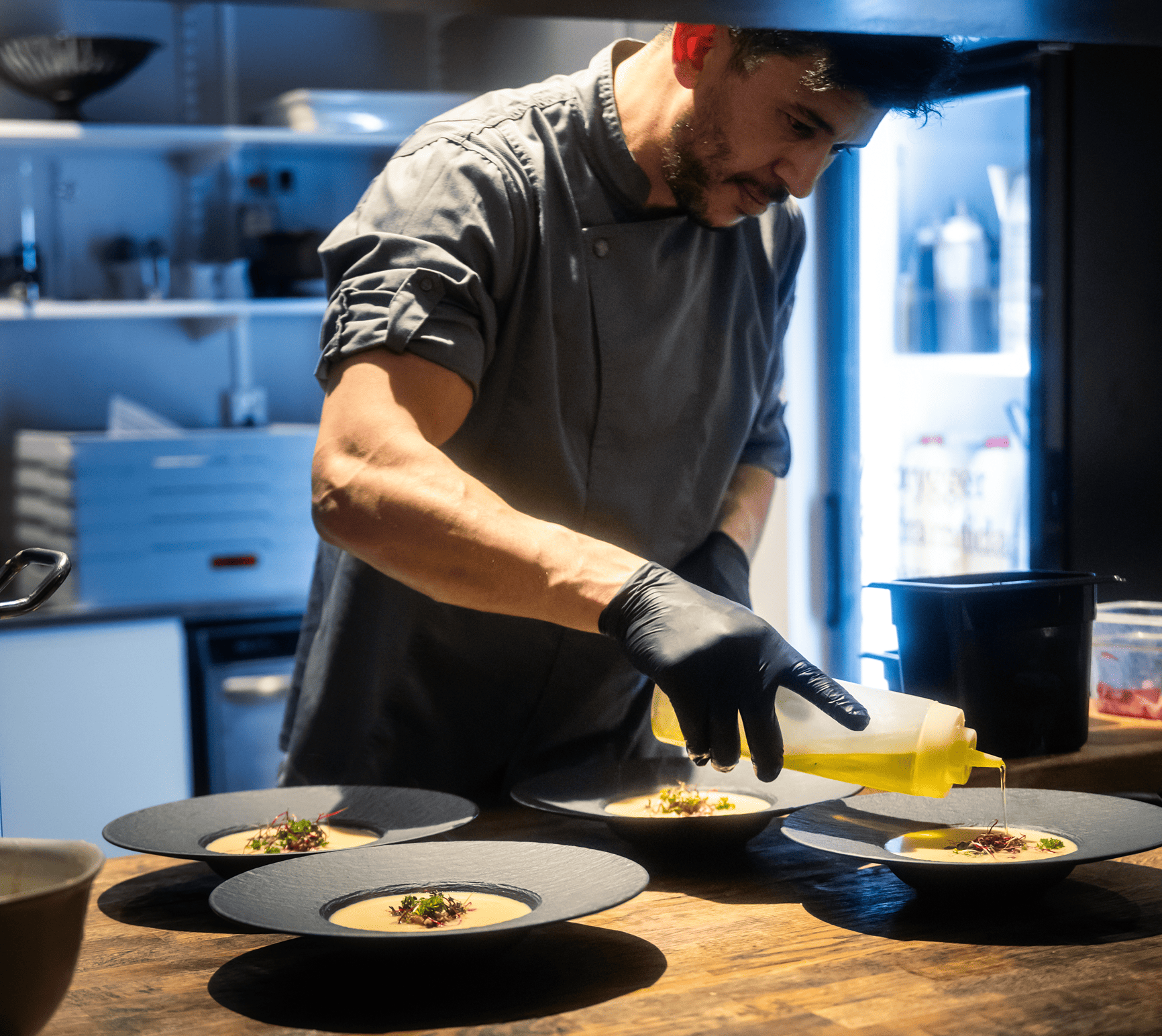 Koch in grauer Kleidung und schwarzen Handschuhen gibt Öl auf Teller mit Suppe und Garnitur in einer professionellen Küche.