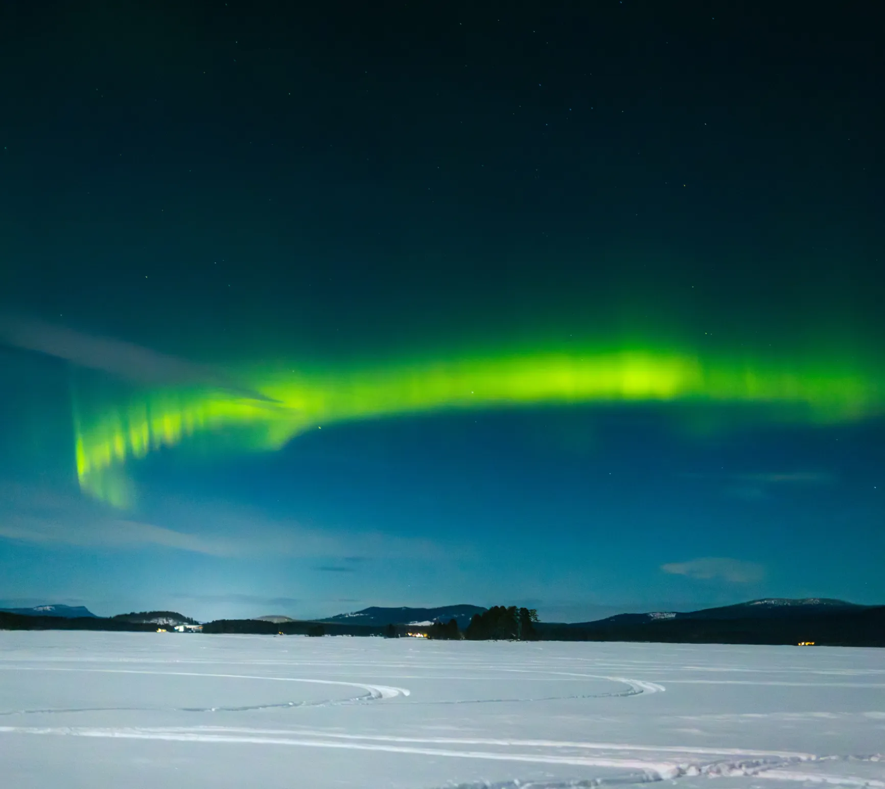 Leuchtende grüne Nordlichter am Nachthimmel über verschneiter Landschaft mit Bergen und einigen Lichtern am Horizont.