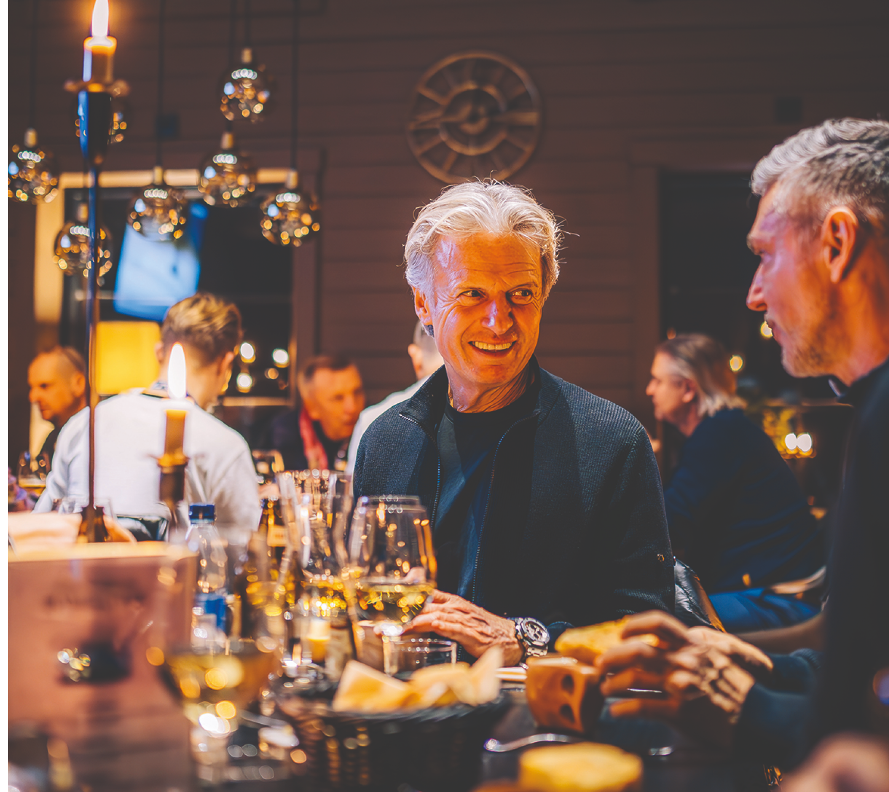 Älterer Mann mit grauen Haaren sitzt an einem Tisch mit Weingläsern und isst Brot in einem gemütlich beleuchteten Restaurant.