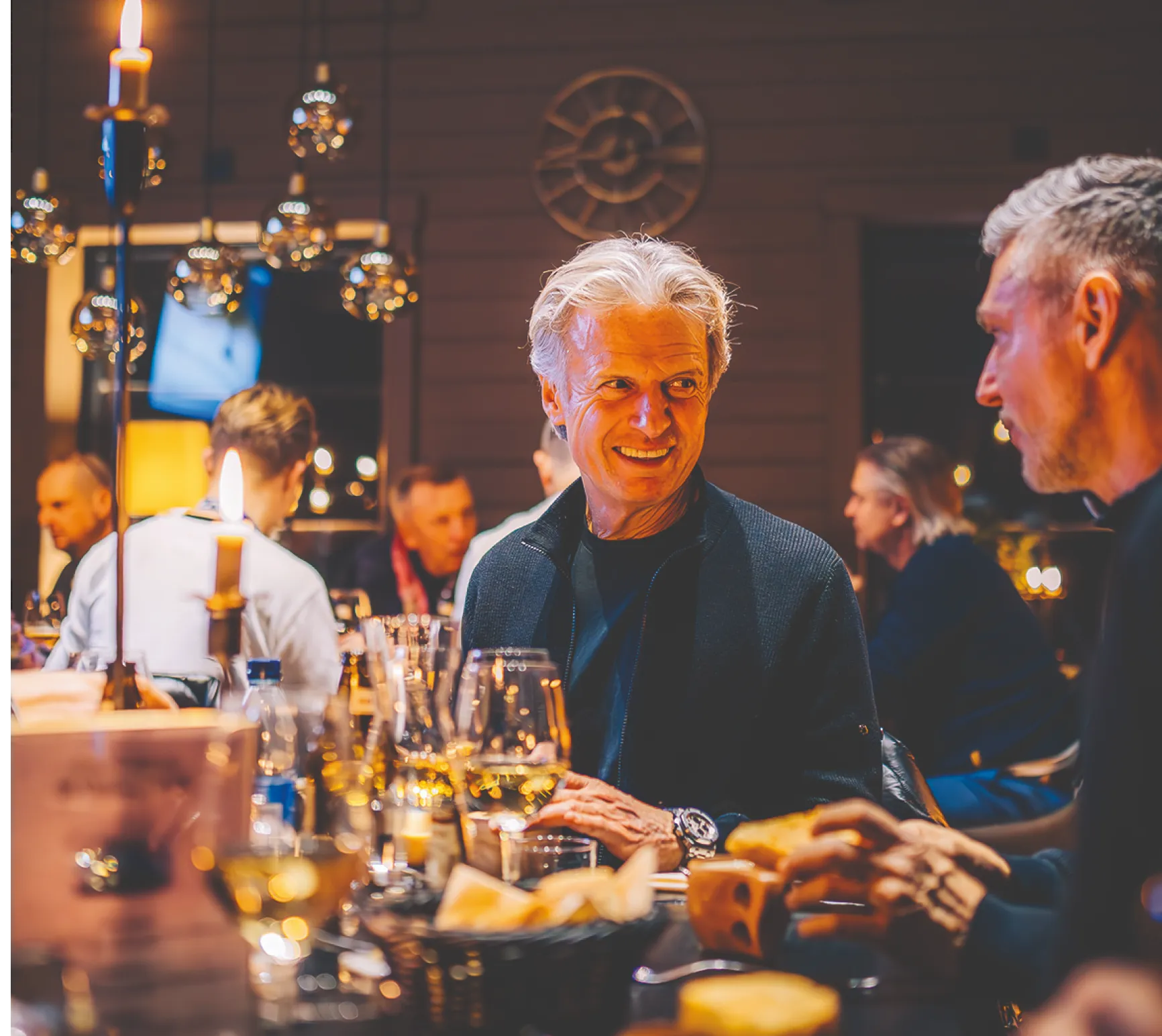 Älterer Mann mit grauen Haaren sitzt an einem Tisch mit Weingläsern und isst Brot in einem gemütlich beleuchteten Restaurant.