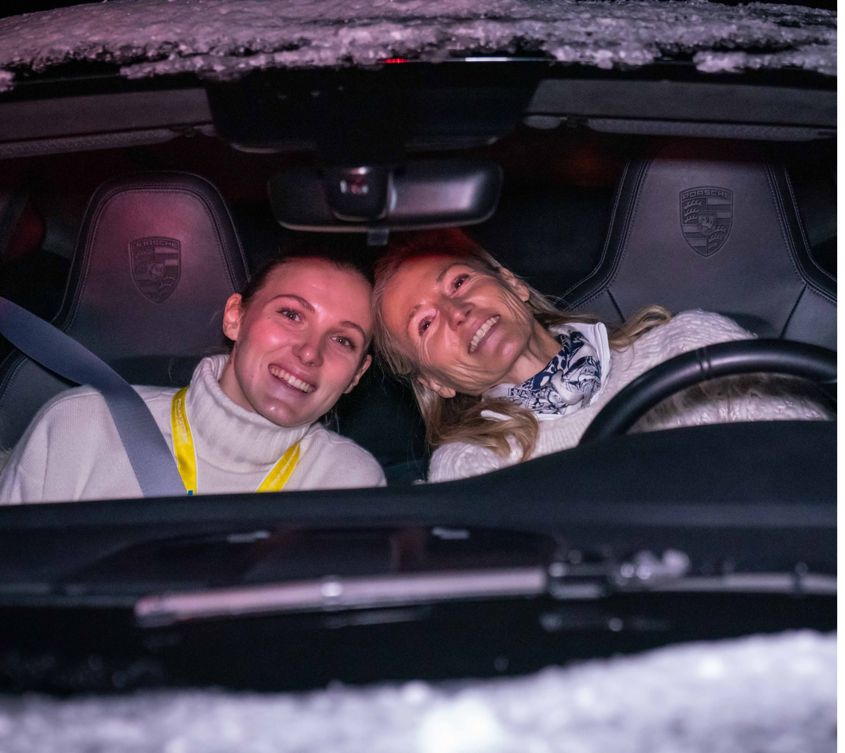 Zwei lächelnde Frauen sitzen vorne im Porsche bei Nacht, beide tragen weiße Pullover und sind angeschnallt.
