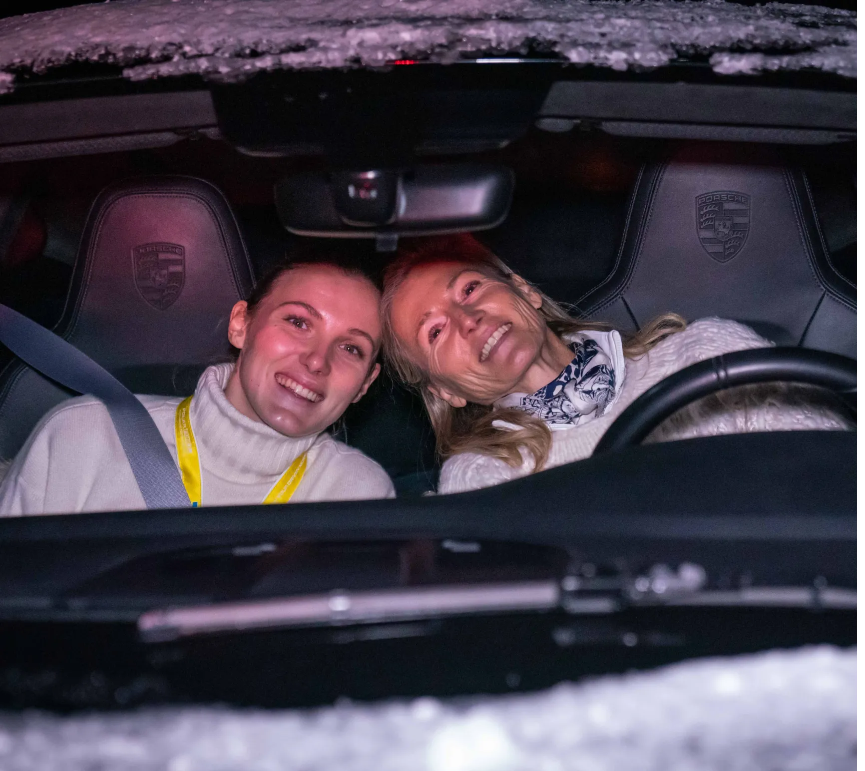 Zwei lächelnde Frauen sitzen vorne im Porsche bei Nacht, beide tragen weiße Pullover und sind angeschnallt.