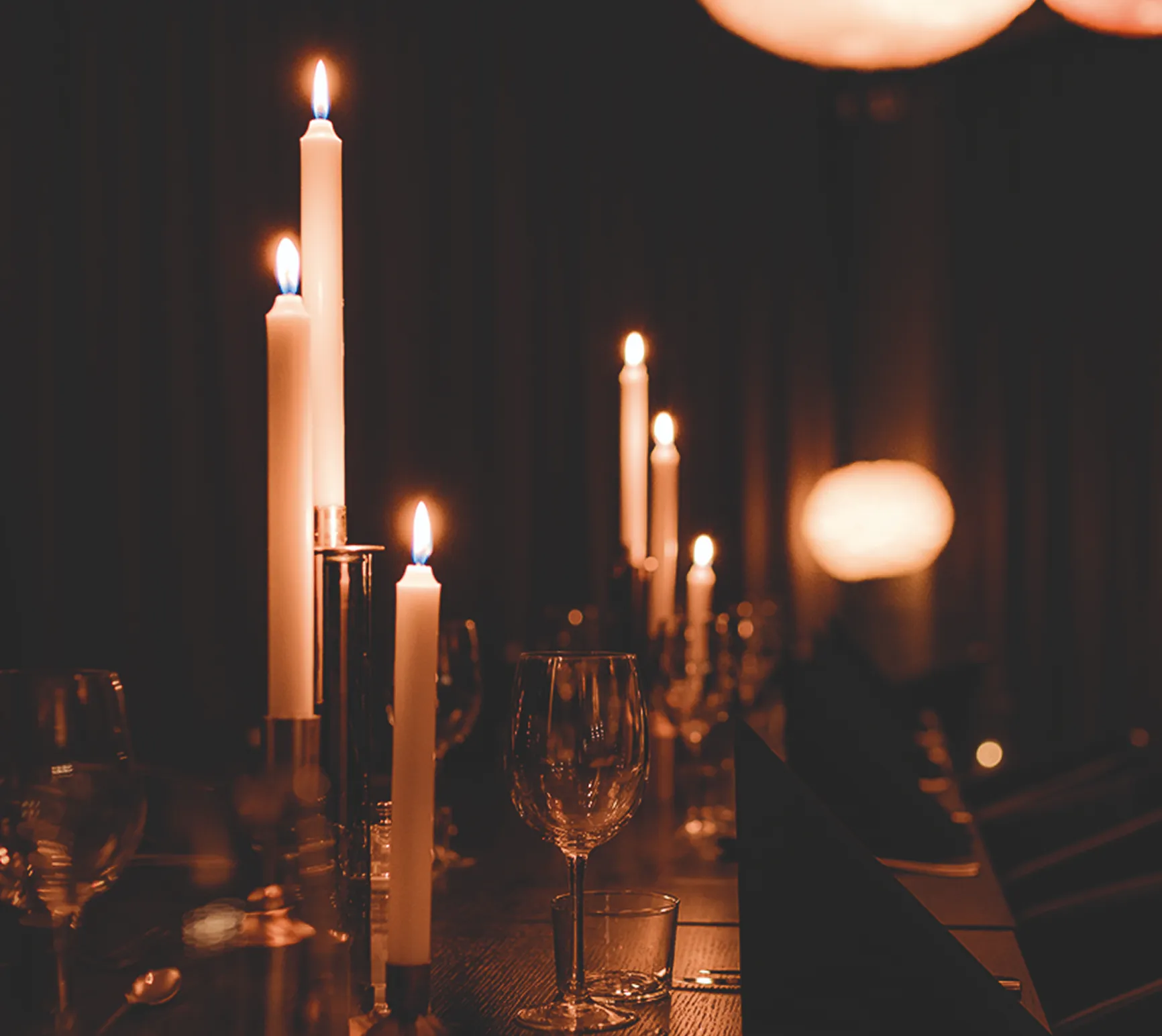 Kerzen in Kerzenhaltern auf einem festlich gedeckten Tisch mit Weingläsern in gedämpftem Licht.
