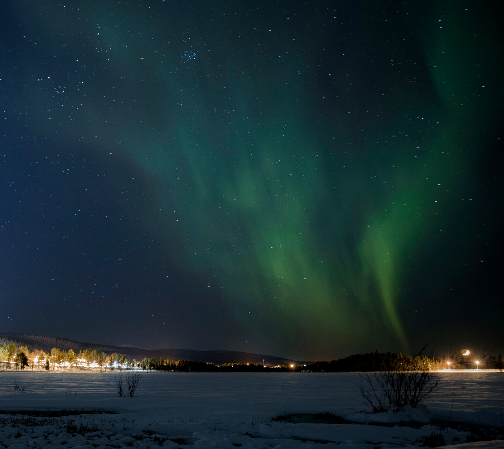 Nordlichter über einer schneebedeckten Landschaft mit Sternenhimmel und beleuchteten Bäumen am Horizont.