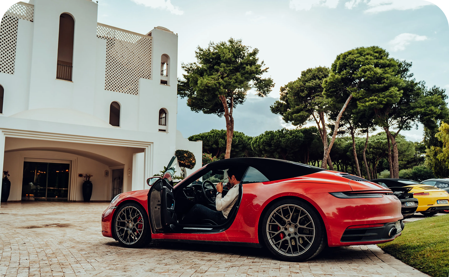 Roter Sportwagen mit geöffnetem Tür, daneben sitzt ein Mann, im Hintergrund weiße Villa und Bäume.