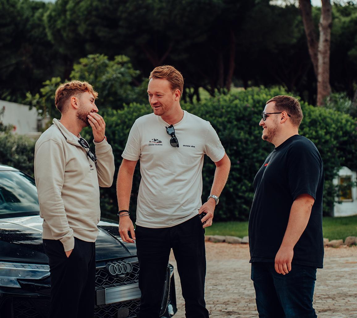 Drei Männer stehen vor einem schwarzen Audi und unterhalten sich im Freien, umgeben von grünen Büschen und Bäumen.