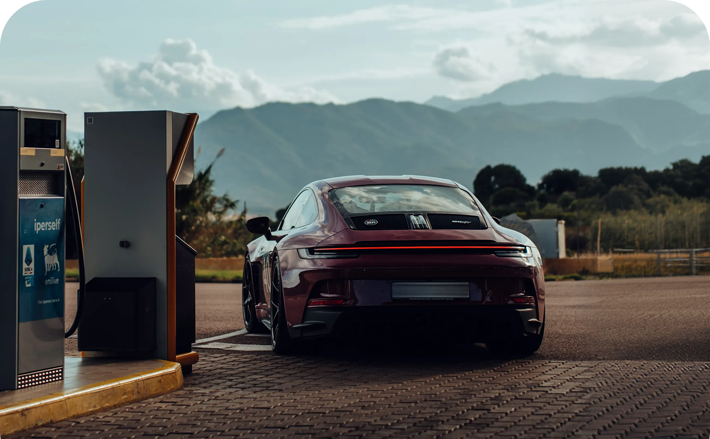 Rotes Sportauto an einer Tankstelle mit Bergen und bewölktem Himmel im Hintergrund.