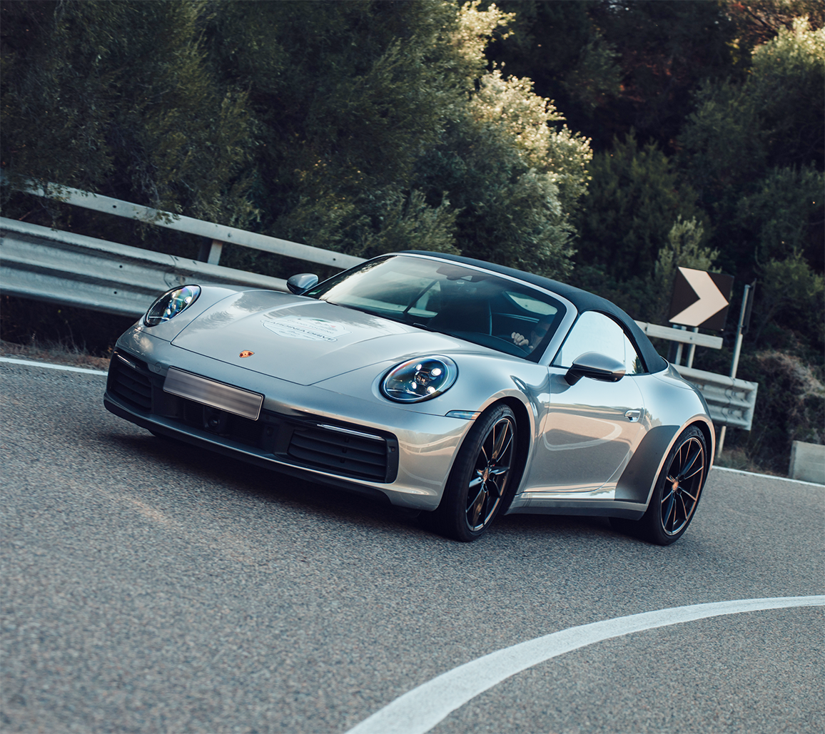 Silberner Porsche-Sportwagen mit schwarzem Verdeck fährt auf kurviger Straße mit Wald im Hintergrund.