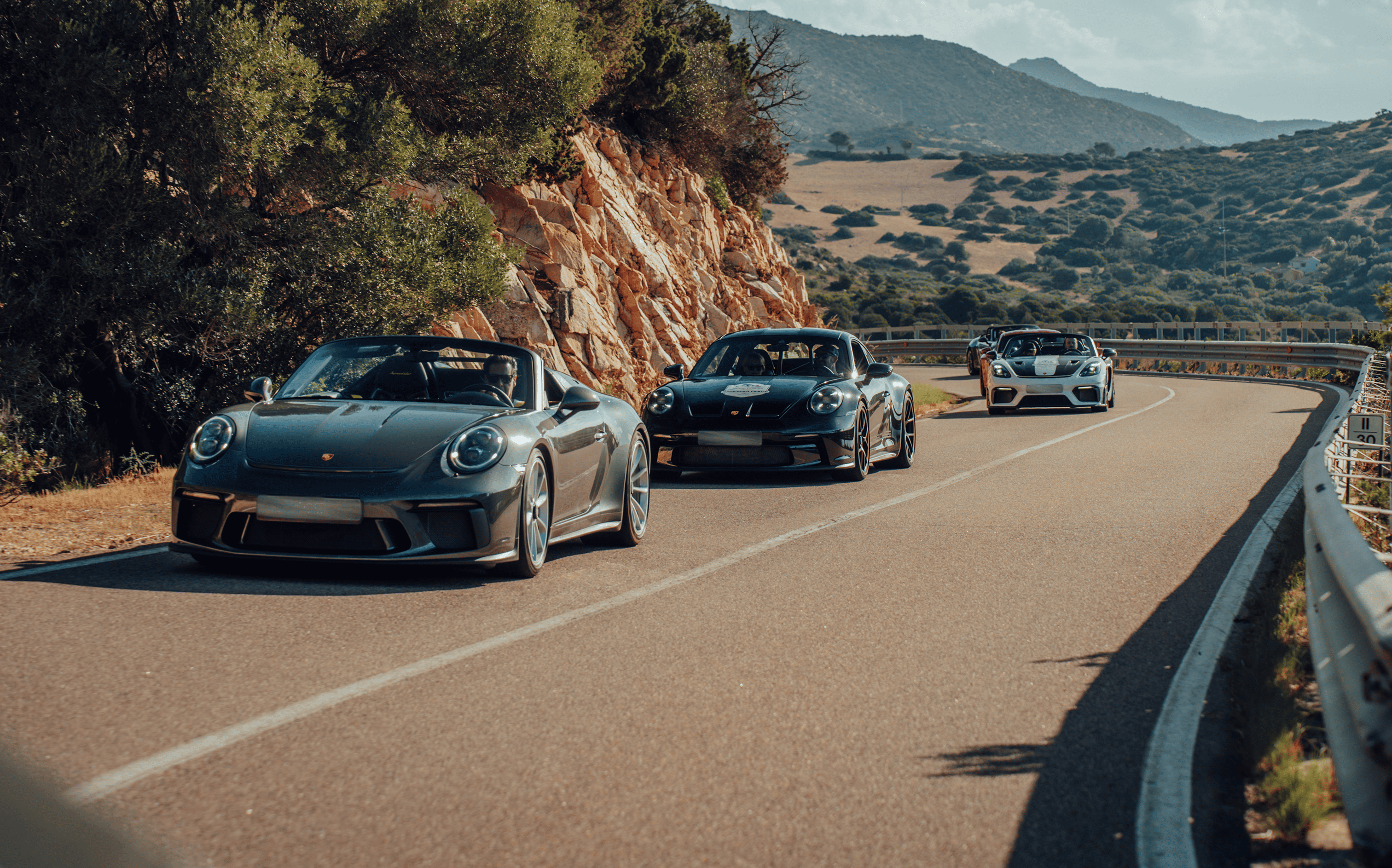 Drei Porsche-Sportwagen fahren auf einer kurvigen Landstraße durch eine bergige Landschaft.